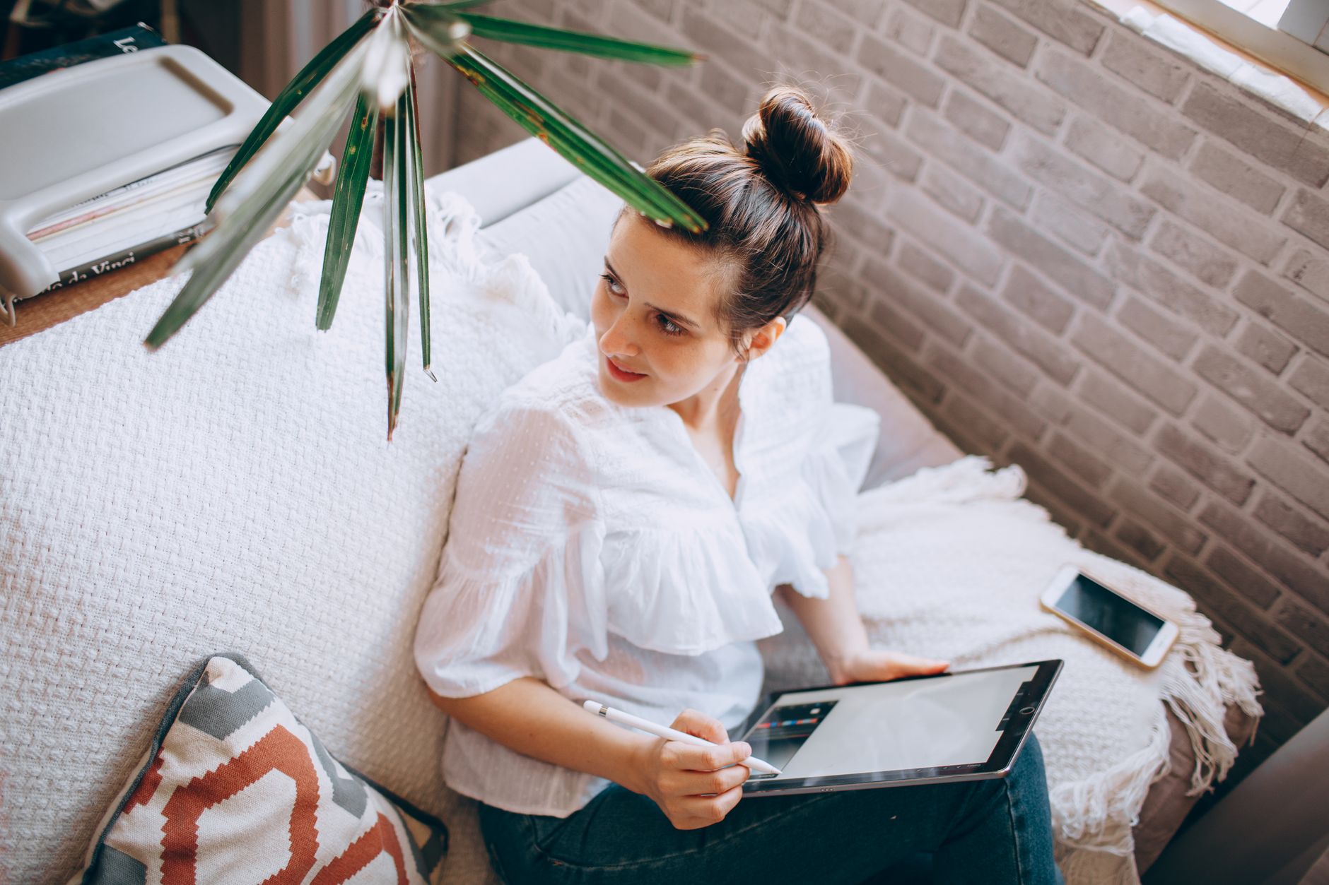 From above of glad female using tablet and stylus while surfing internet on sofa with smartphone
