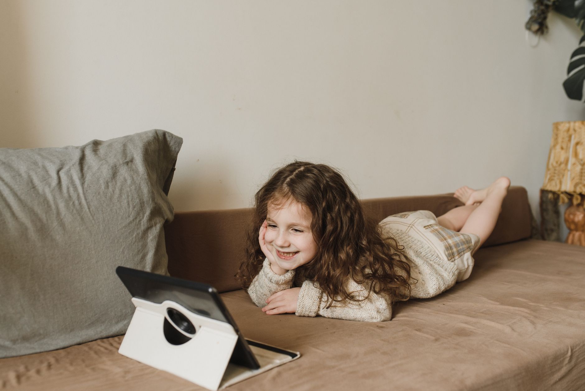 A young girl happily watching a tablet while lying on a couch indoors, exuding warmth and comfort.