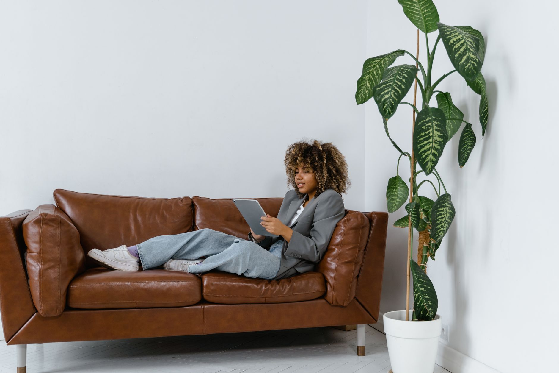 African American woman lounging on a brown leather sofa with a tablet indoors, next to a potted plant.
